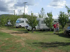 Zwei Wohnmobile auf dem Campingplatz, umgeben von Bäumen und Rasen, im Hintergrund Hügel und bewölkter Himmel.