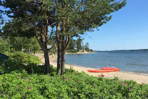 Roter Kajak am Sandstrand, dahinter Bäume und ein Uferweg am See mit weiteren Booten und Liegeplätzen.