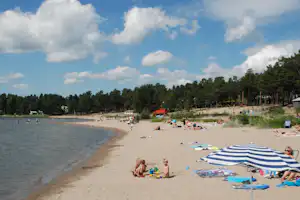 Sandstrand am See mit Badegästen, Liegestühlen und Sonnenschirm, im Hintergrund Wald und ein Spielbereich.