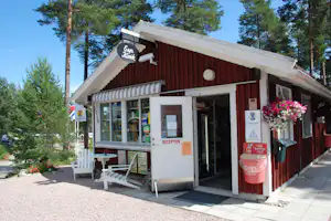 Rezeption eines Campinggebäudes mit rotem Holzvordach, Eingangstür, Sitzbank und Blumen vor Kiefern im Hintergrund.