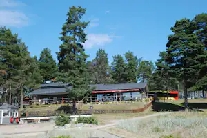 Restaurant mit Terrasse und Sitzplätzen im Grünen, umgeben von hohen Kiefern und einem Bus am Rand des Campingplatzes.