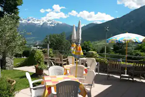 Terrasse mit Tisch und Stühlen, Sonnenschirm und Blick auf grüne Berge mit schneebedeckten Gipfeln.