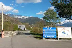 Camping-Einfahrt mit Wohnmobil am Parkplatz, Hinweisschild zur Rezeption und Blick auf die Alpen im Hintergrund.
