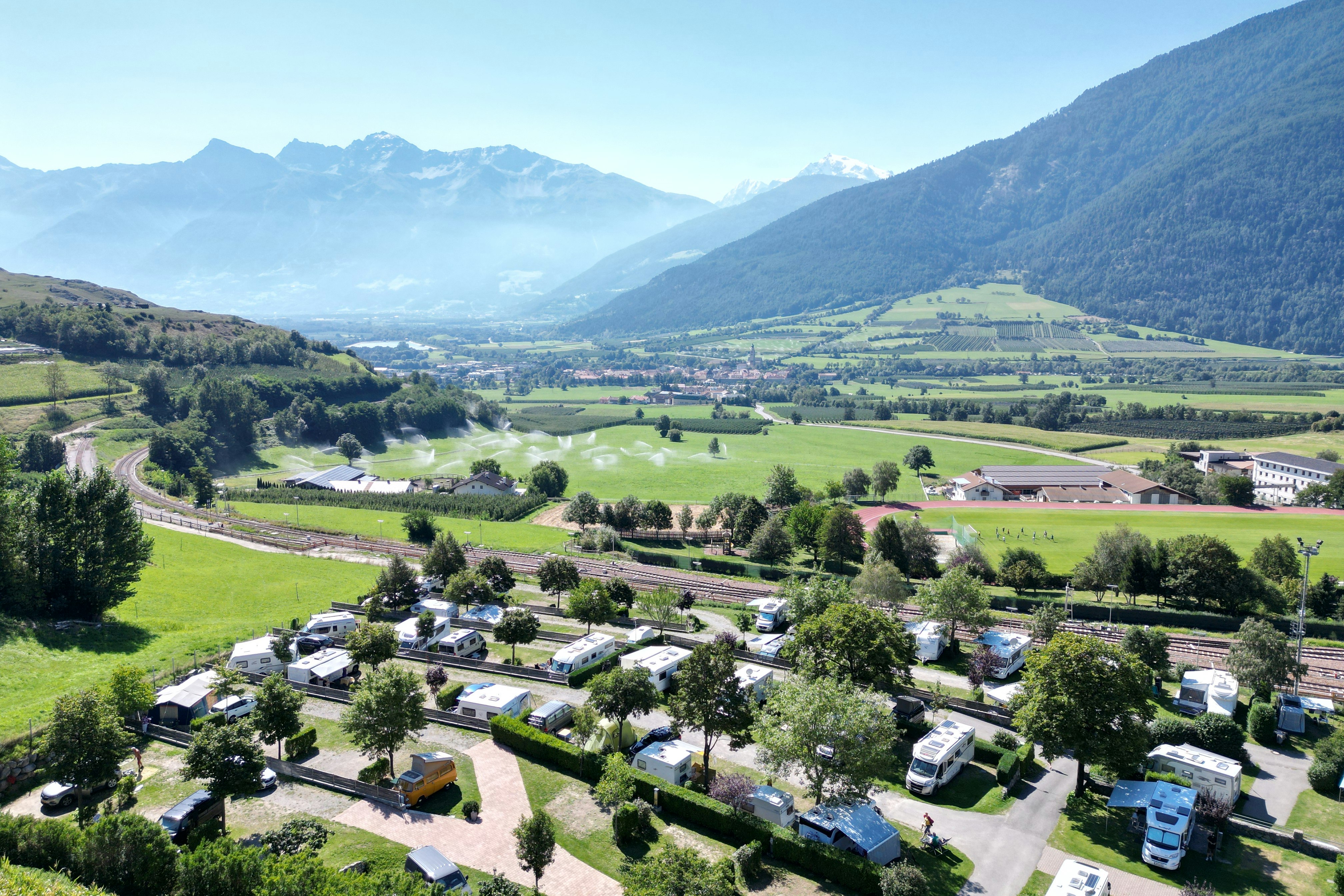 Luftaufnahme eines Campingplatzes mit Wohnwagen und Stellplätzen, gepflegten Wegen, Gärten und Blick ins grüne Tal mit Bergen.
