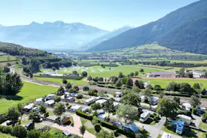 Luftaufnahme eines Campingplatzes mit Wohnwagen und Stellplätzen, gepflegten Wegen, Gärten und Blick ins grüne Tal mit Bergen.