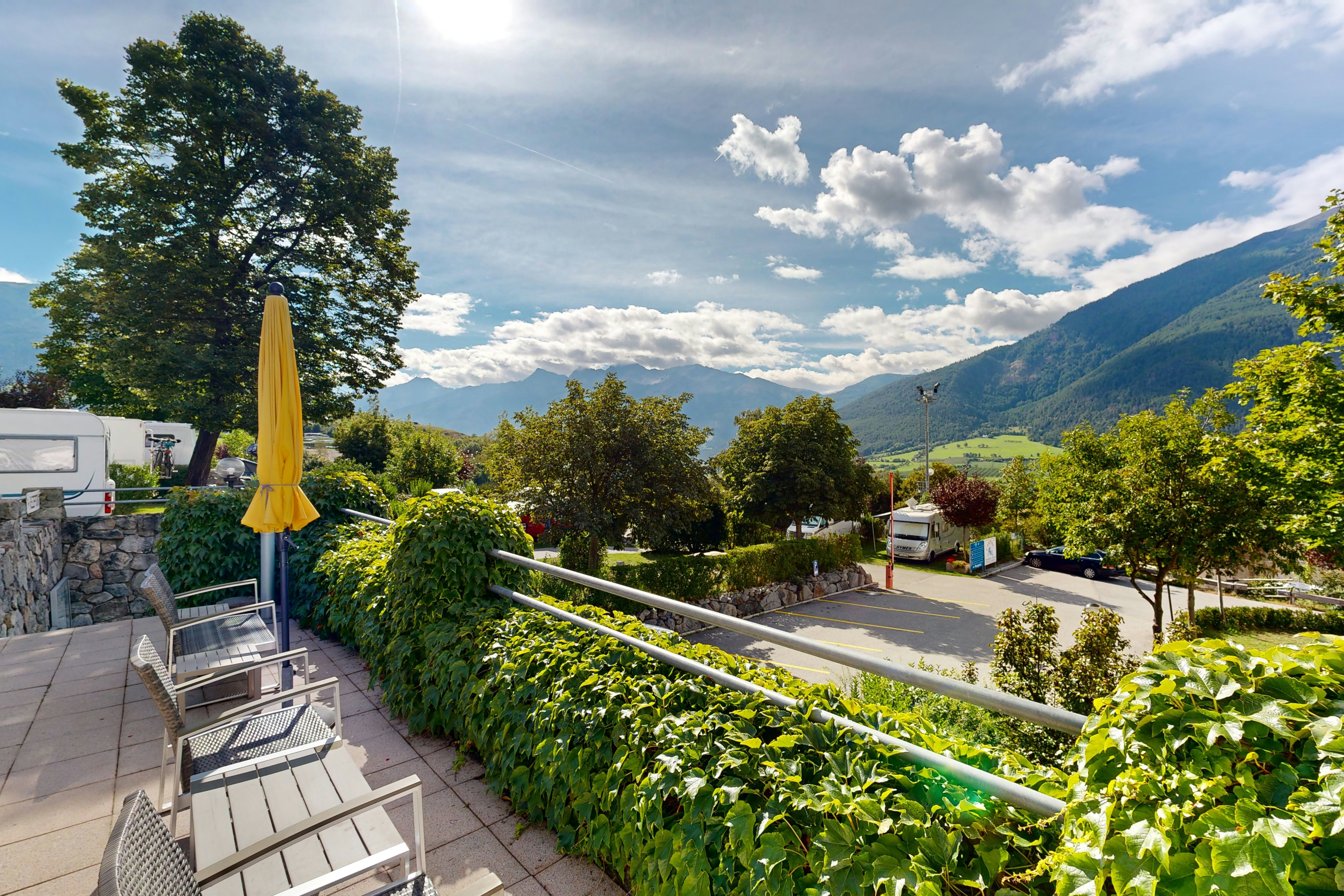 Blick von der Terrasse auf einen Campingplatz mit Hecken, Sitzbank und Liegestühlen, im Hintergrund Berge und Bäume.
