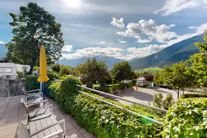 Blick von der Terrasse auf einen Campingplatz mit Hecken, Sitzbank und Liegestühlen, im Hintergrund Berge und Bäume.