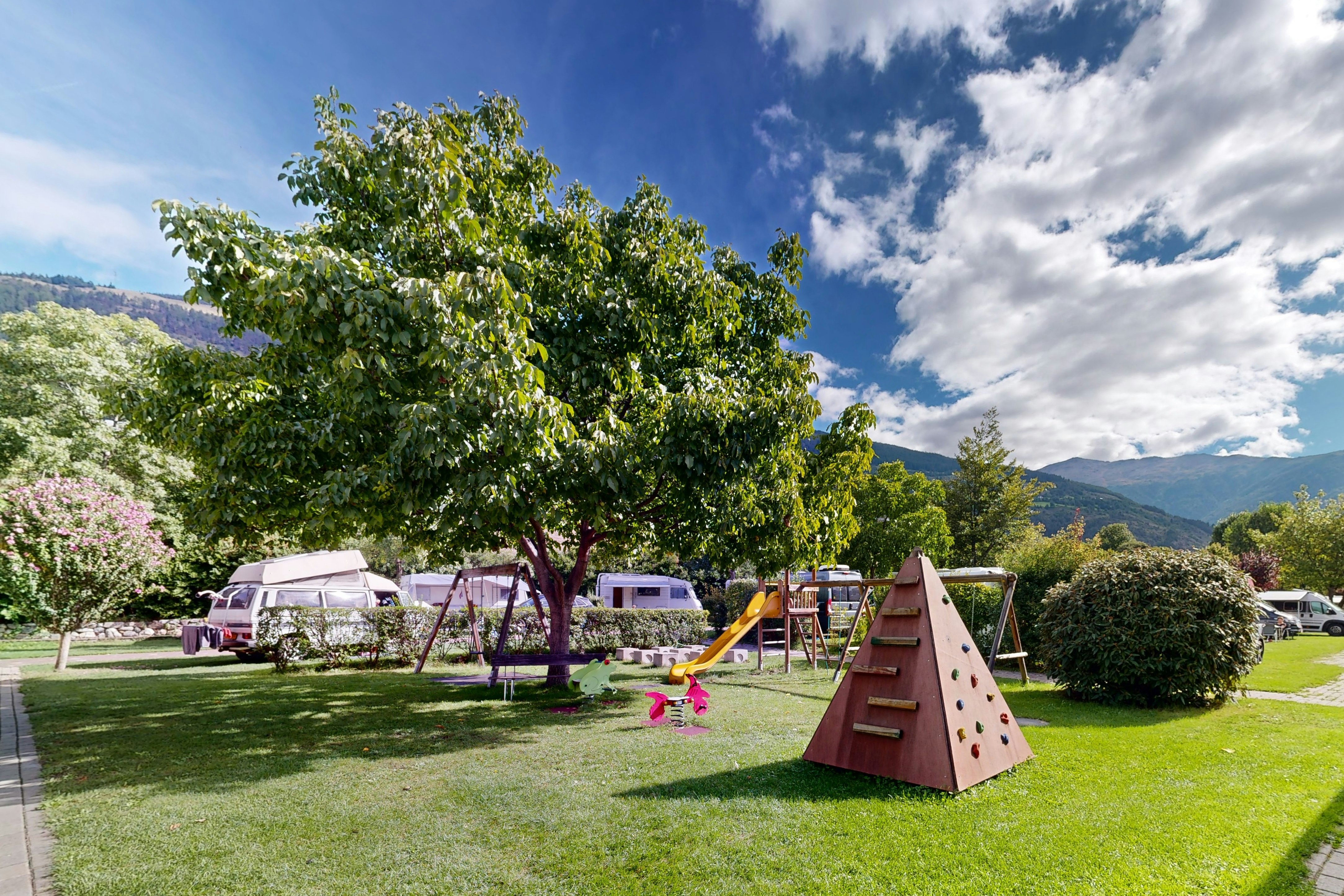 Spielbereich im Campinggarten mit Rutsche, Schaukel und Rasenfläche, daneben Wohnwagen und Bäume vor Bergen und blauem Himmel.