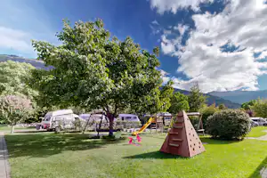 Spielbereich im Campinggarten mit Rutsche, Schaukel und Rasenfläche, daneben Wohnwagen und Bäume vor Bergen und blauem Himmel.