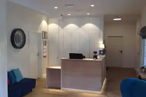 Rezeption mit Holztheke, blauen Sesseln und Wanduhren in einer hellen Camping-Lobby mit Flur und Türen.
