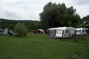 Wohnwagen und Zelt auf grünem Campingplatz, umgeben von Bäumen und Hügeln im Hintergrund.