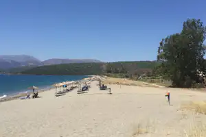 Sandstrand mit Sonnenschirmen und Liegen am Ufer, im Hintergrund bewaldete Hügel und blaues Wasser.