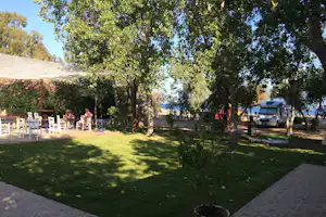 Campinggarten mit Rasen, Bäumen und Sitzgruppe unter einem großen Sonnensegel, im Hintergrund Wohnmobil und Weg zum Meer.