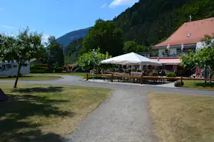 Terrasse mit großen Sonnenschirmen, Holztischen und Stühlen am Campingplatz, im Hintergrund Berge und dichte Bäume.