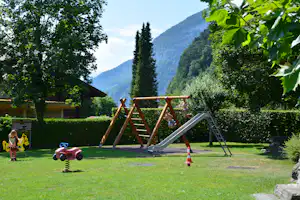 Spielplatz mit Holzrutsche und Klettergerüst im grünen Garten, Kinderfahrzeug und Berge im Hintergrund.