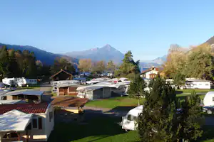 Campingplatz mit Wohnwagen und Mobilheimen, Blick auf Berge und bewaldete Hänge, im Vordergrund Rasen und Bäume.