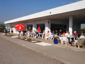 Terrasse am Campingplatz mit überdachtem Restaurant, vielen weißen Stühlen und Tischen sowie rotem Sonnenschirm vor dem Gebäude.