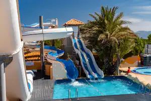 Camping-Pool mit blauen Wasserrutschen, Palmen und Liegen, im Hintergrund ein Gebäude mit Terrassendach.