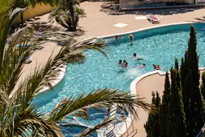 Großer Camping-Pool mit Sonnendeck, Palmen und Liegen; im Hintergrund Wege, Hecken und ein umzäunter Bereich.