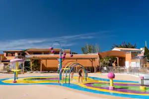 Buntes Kinderbecken mit Wasserrutsche und Spielbogen, daneben Terrassen und Gebäude am Campingplatz unter blauem Himmel.