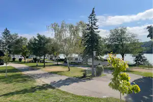 Campingplatz mit Wohnmobilen und Bäumen am See, gepflasterter Weg und Rasenflächen zwischen den Stellplätzen.