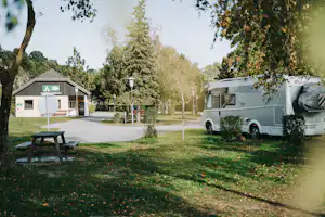 Wohnwagen auf dem Campingplatz neben einem kleinen Gebäude, gepflasterter Weg und Rasen mit Bäumen im Hintergrund.