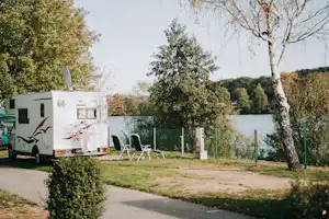 Wohnwagen auf einem Campingplatz am Flussufer, mit Stühlen und Tisch am Stellplatz, umgeben von Bäumen und Grün.