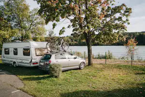 Wohnmobil und Pkw am Stellplatz am See, mit Bäumen, Grasfläche und Zaun im Hintergrund.