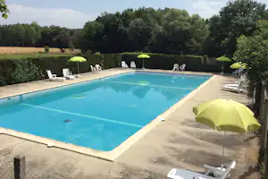 Blauer Camping-Pool mit Betonrand, grünen Sonnenschirmen und weißen Liegestühlen auf der Terrasse, umgeben von Hecken und Bäumen.
