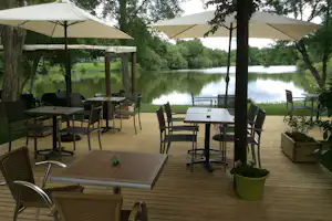 Terrasse am See mit Holzboden, Tischen und Stühlen unter Sonnenschirmen, im Hintergrund Bäume und ruhiges Wasser.