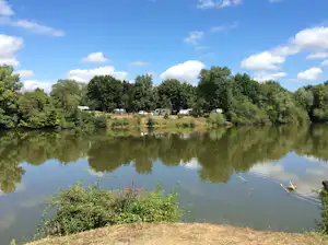 Ruhiger See am Campingplatz mit Uferböschung, Bäumen und Wohnwagen am Rand, reflektiert im Wasser.