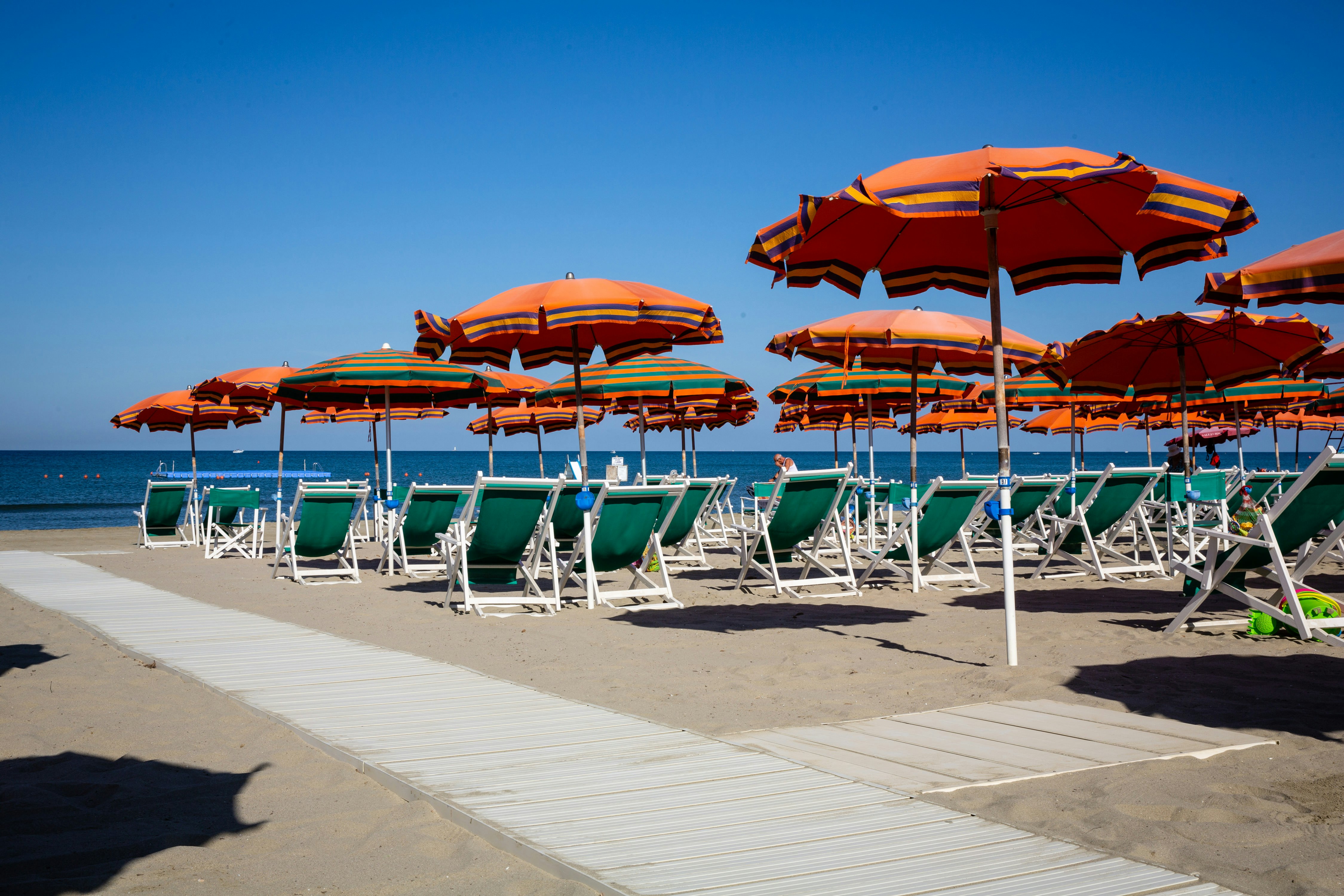 Strand mit bunten Sonnenschirmen und Liegen auf einem Campingplatz, daneben ein gepflasterter Weg und das Meer.