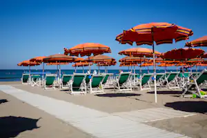 Strand mit bunten Sonnenschirmen und Liegen auf einem Campingplatz, daneben ein gepflasterter Weg und das Meer.