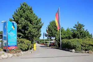 Camping-Eingang mit Schildern, deutschen Flaggen und breitem Weg zwischen Bäumen und Hecken.
