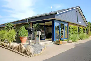 Campinghaus mit überdachter Terrasse und großen Glasfronten, gepflasterter Weg, Hecken und Steine im Garten.