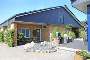 Bungalow mit Ziegelwand, Eingangstür und Fenstern, gepflasterte Terrasse mit Sitzmöbeln, Pflanzen und Kiesbeet im Vordergrund.