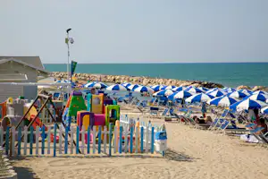 Strand mit Campingbereich, blauen Sonnenschirmen und Liegestühlen, weißem Zaun und Spielzeug am Sand.
