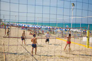 Strandvolleyballfeld im Campingbereich mit Netz, Sand und Spielern, im Hintergrund ein blaues Wasserbecken und Sonnenschirme.