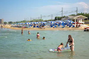 Badegäste im Meer vor einem Campingstrand mit vielen blauen Sonnenschirmen und Liegen, im Hintergrund ein Gebäude.