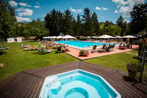 Blick auf den Pool mit Whirlpool auf der Terrasse, Liegestühlen und Sonnenschirmen am Becken im Grünen.