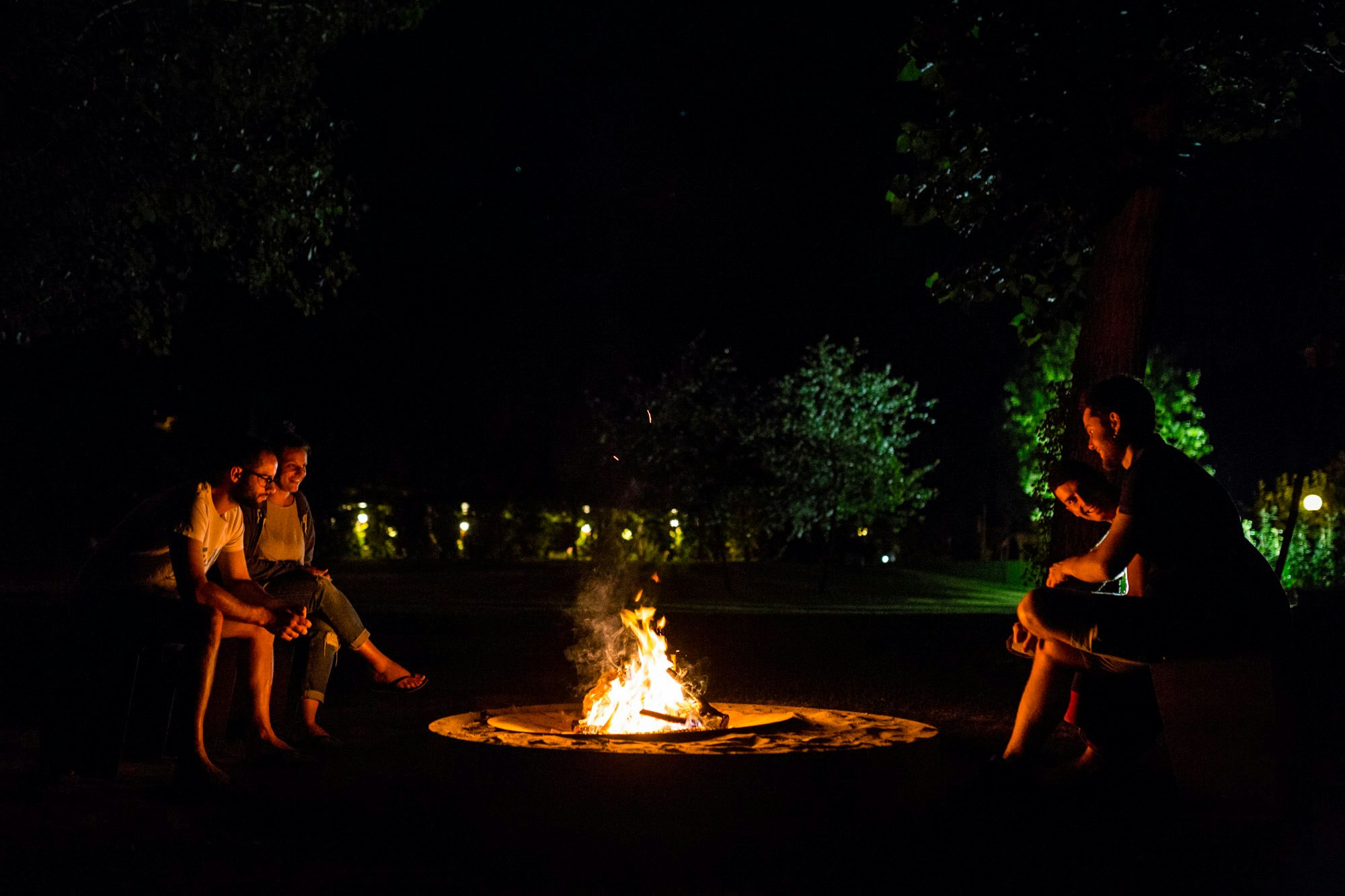 Zwei Personen sitzen abends am Lagerfeuer auf einem Campingplatz, im Hintergrund sind Bäume und beleuchtete Wege zu sehen.