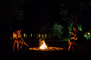Zwei Personen sitzen abends am Lagerfeuer auf einem Campingplatz, im Hintergrund sind Bäume und beleuchtete Wege zu sehen.