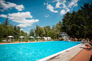 Campingpool mit blauem Wasser, Holzterrasse, Liegen und Sonnenschirmen, im Hintergrund Bäume und ein Spielbereich.