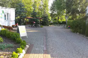 Gepflasterter Weg am Campingplatz mit Schotterfläche, Fahrradständer und Bäumen neben einem weißen Gebäude.
