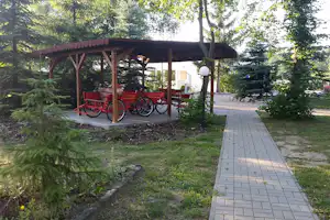 Überdachter Unterstand mit roten Fahrrädern unter Holzpfeilern, gepflasterter Weg und Bäume im Campinggarten.