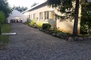 Campingplatz-Weg am Wohngebäude mit Terrasse, Zelt und Holztisch, umgeben von Büschen und Bäumen.