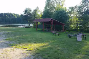 Holzüberdachter Grill- und Sitzbereich mit Bänken auf dem Campingplatz am See, umgeben von Bäumen und Rasen.
