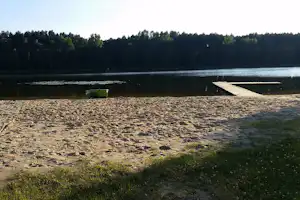 Sandstrand am See mit Steg und Bootssteg, im Hintergrund Wald und ruhiges Wasser.