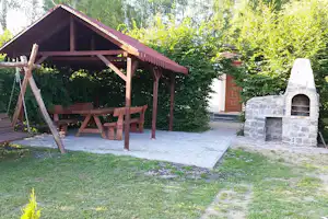Holzunterstand mit Sitzbank und Tisch, daneben ein gemauerter Grill im Garten mit gepflastertem Weg und viel Grün.