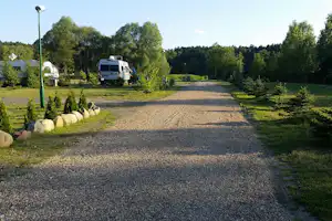 Schotter-Campingstellplatz mit Wohnmobil, Laternen und gepflegten Büschen, umgeben von Bäumen und Rasenflächen.